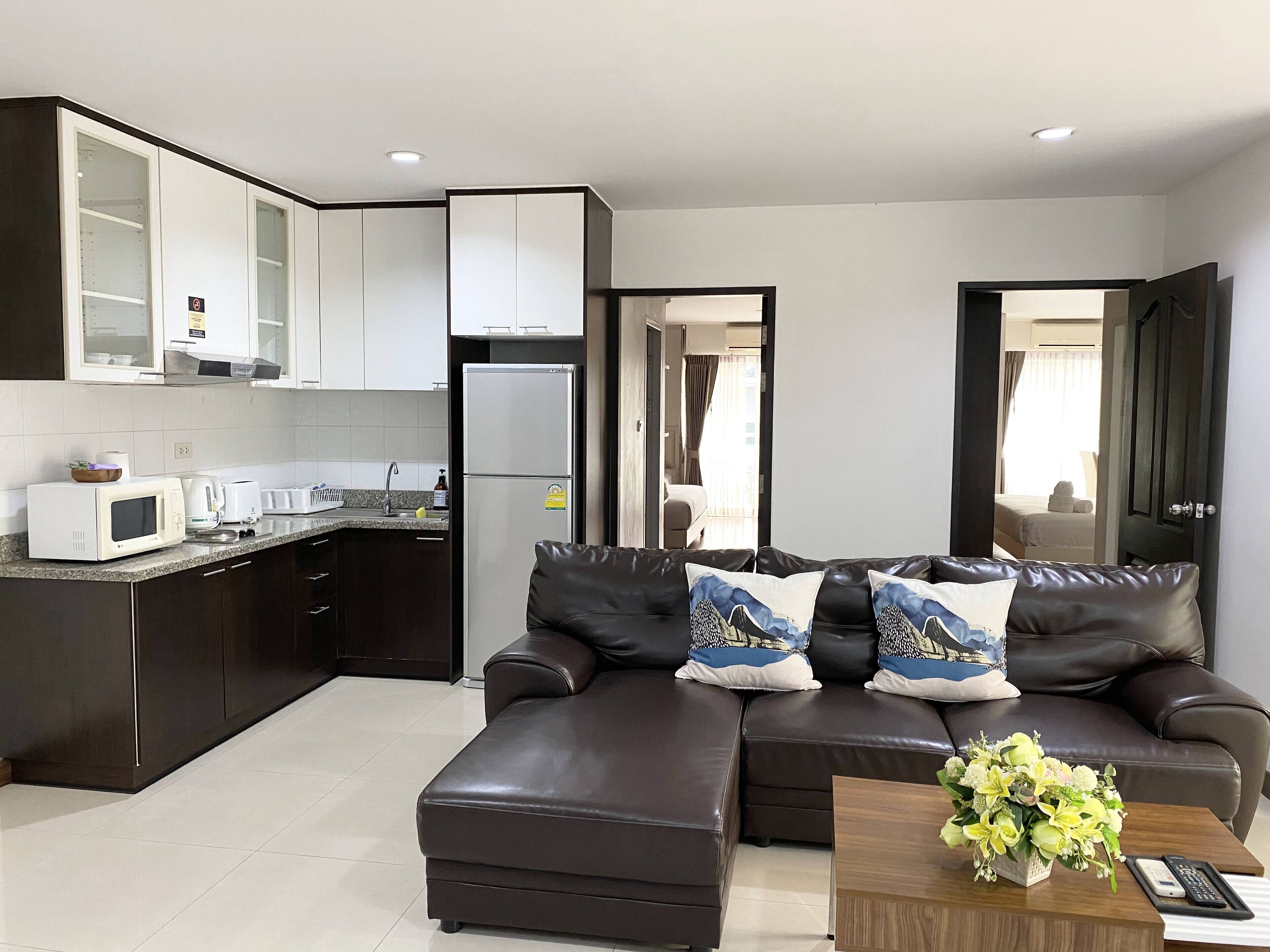 A modern kitchen with dark cabinetry and a white countertop, a brown leather sofa with patterned cushions, and a wooden coffee table with a floral arrangement.