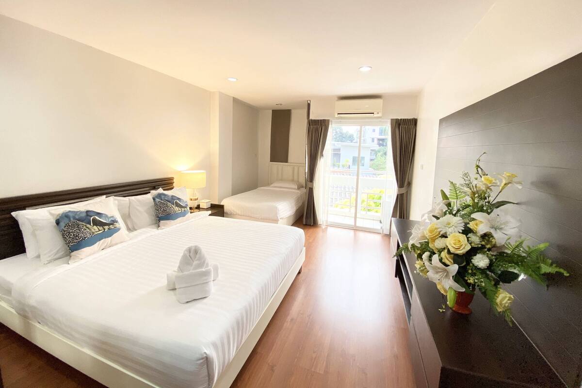 A hotel room with a large bed, a bedside table with a lamp, a small table with a vase of flowers, and a sliding glass door to a balcony.