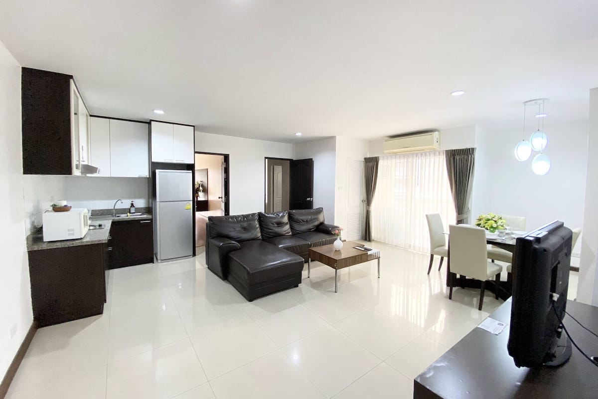 A modern living room with a sofa, coffee table, and TV. There is a kitchen area with cabinets and a refrigerator.