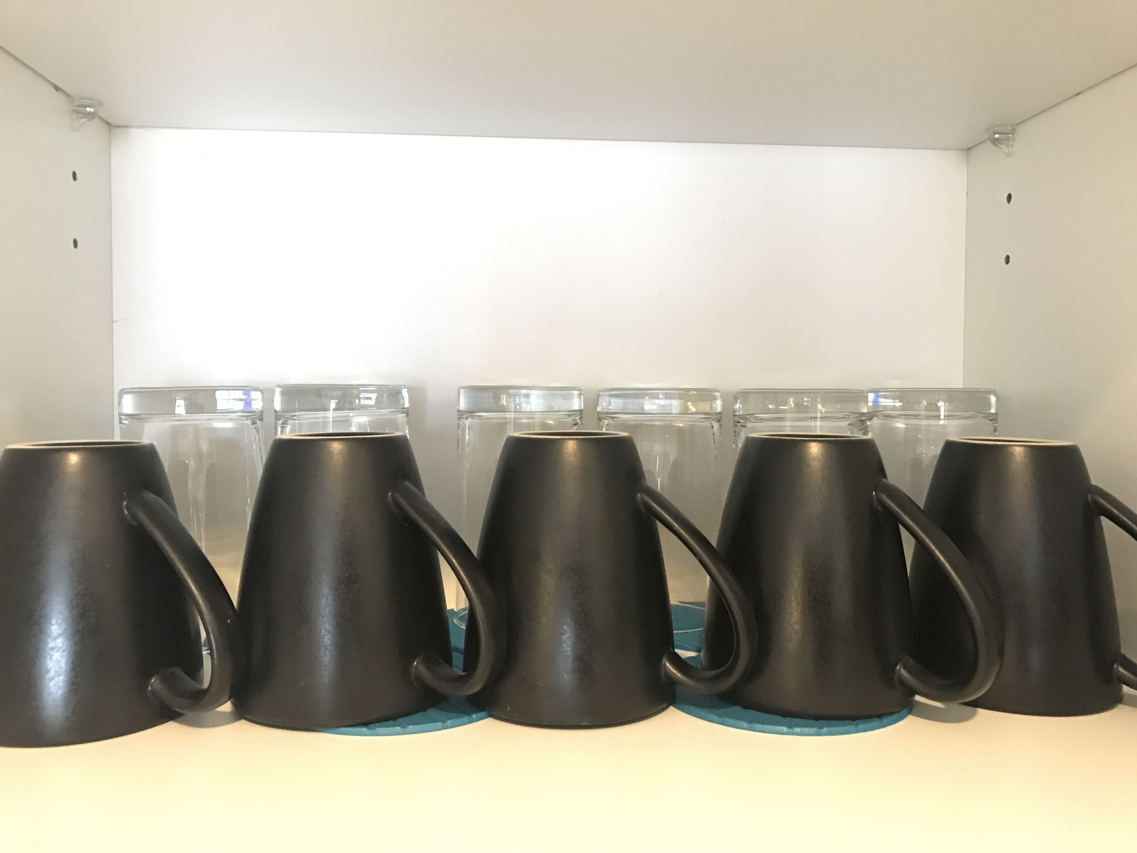 A row of black mugs on a shelf with clear glasses behind them.