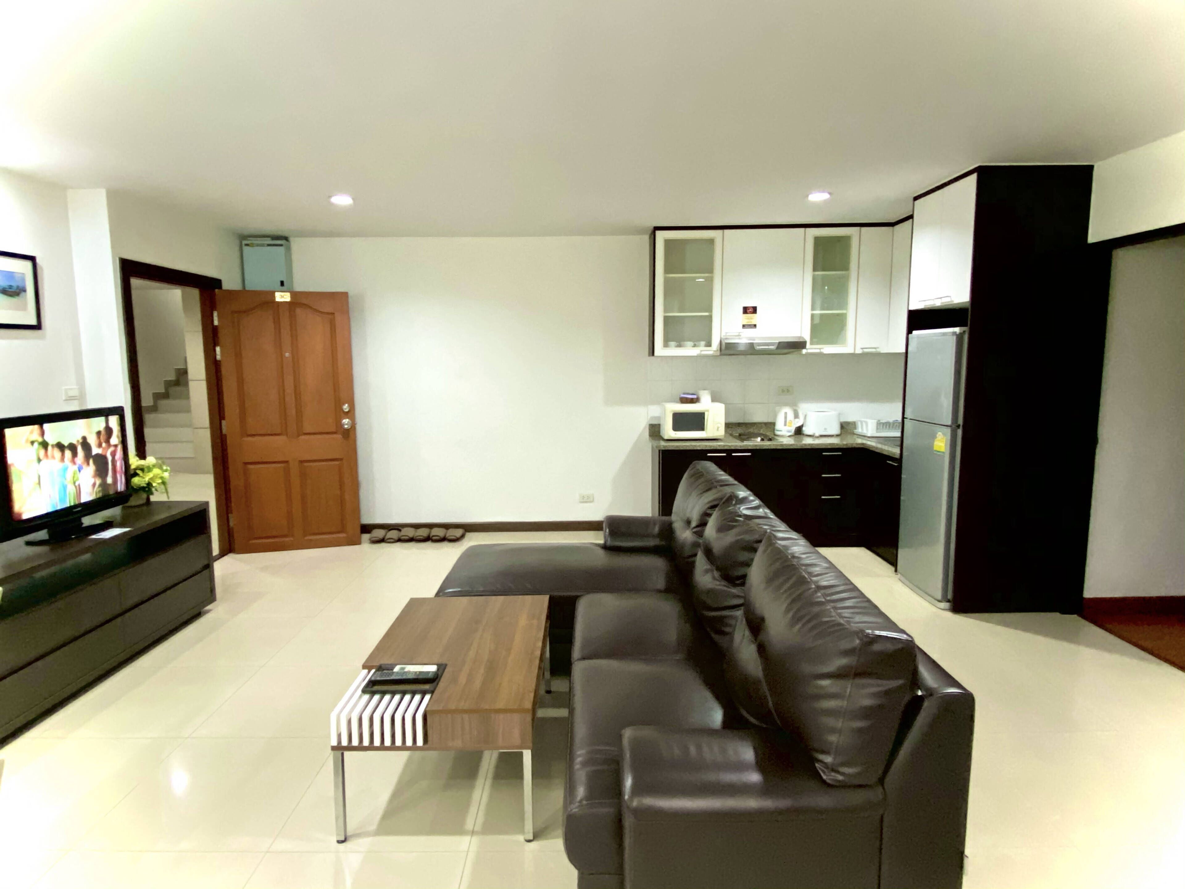 A modern living room with a leather sofa, a wooden coffee table, and a TV on a black stand. A kitchen with a refrigerator, microwave, and cabinets is visible in the background.
