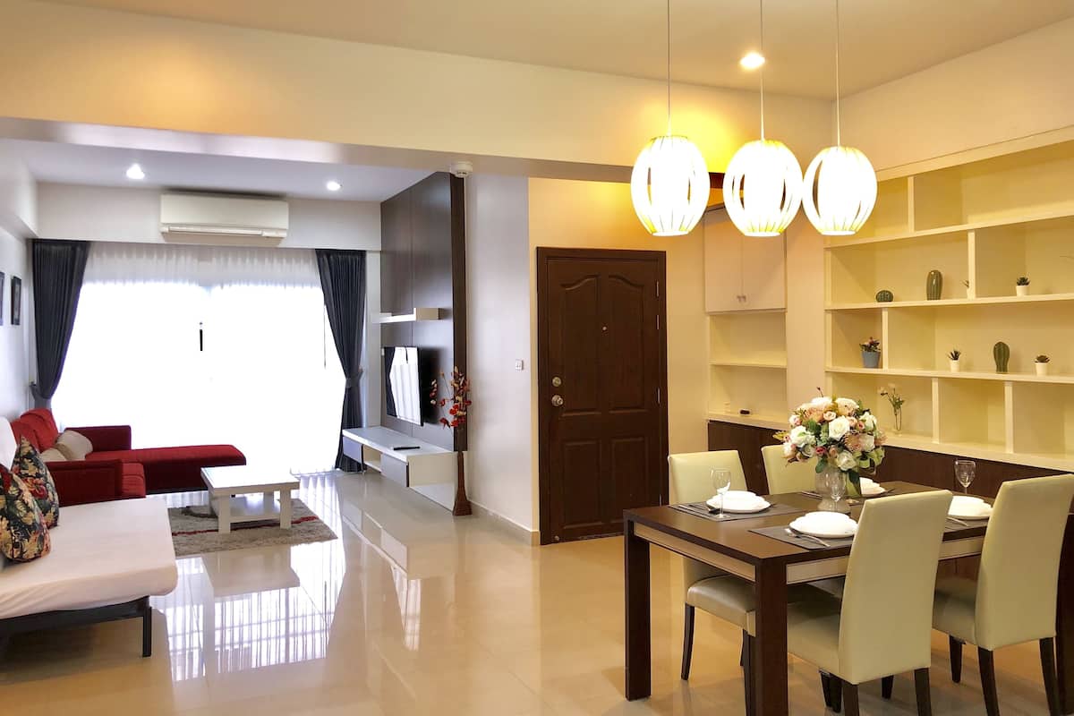 A modern living room with a sofa, dining table, and chairs. There is a TV mounted on the wall, a bookshelf, and a door.