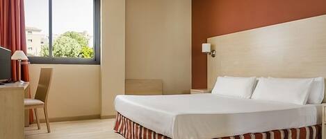 Una habitación de hotel con cama, escritorio, silla, ventana con cortinas y una lámpara en la pared.