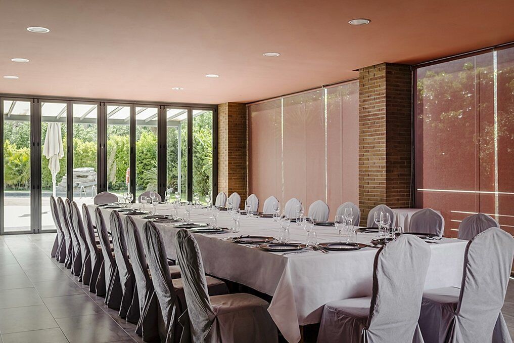 A long dining table set for a formal event, with chairs covered in white fabric, in a room with large windows and a brick wall.