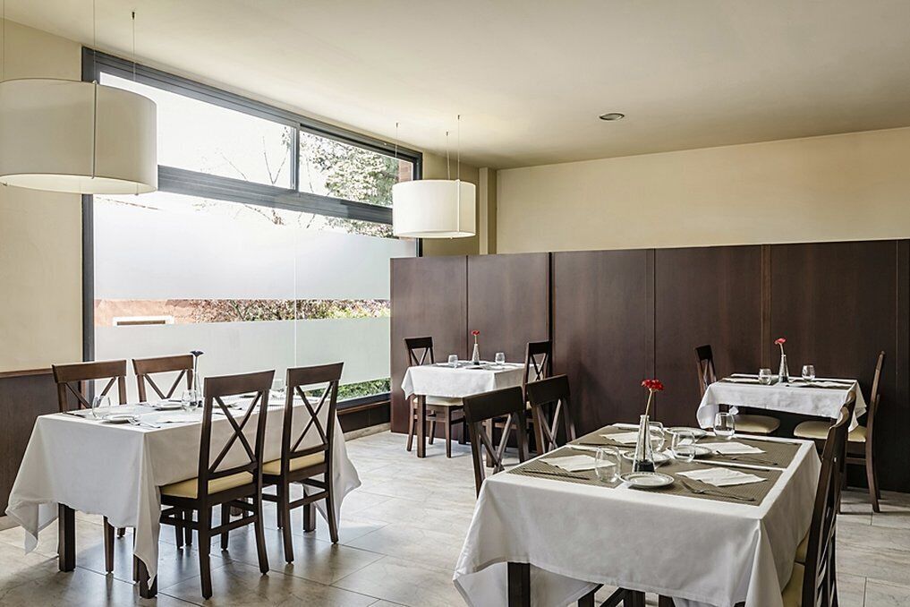 A restaurant with multiple tables set for dining, large windows, and hanging pendant lights.