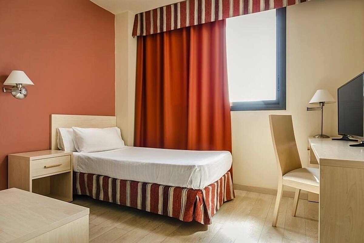 Une chambre d’hôtel avec un lit, un bureau, une chaise, une télévision et une fenêtre avec des rideaux.