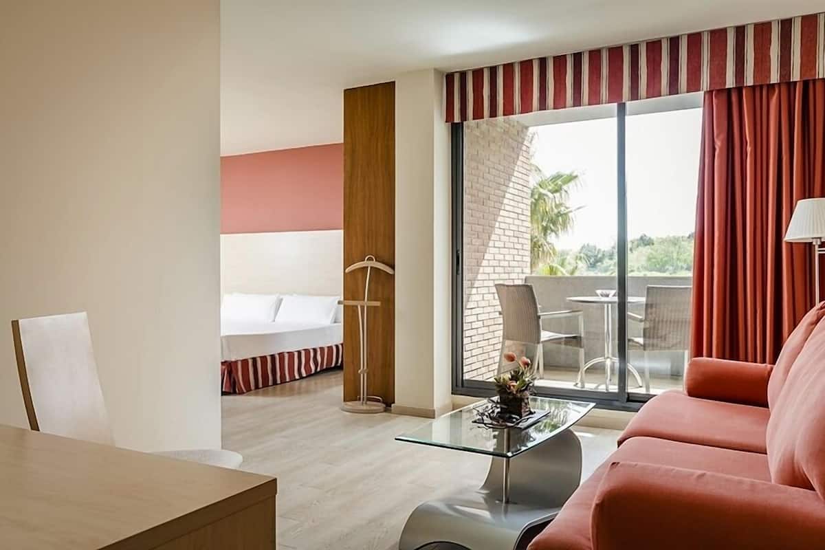 Une chambre d’hôtel avec une table basse en verre, un canapé rouge, un lit avec une literie à rayures et une vue sur l’extérieur grâce à de grandes fenêtres.