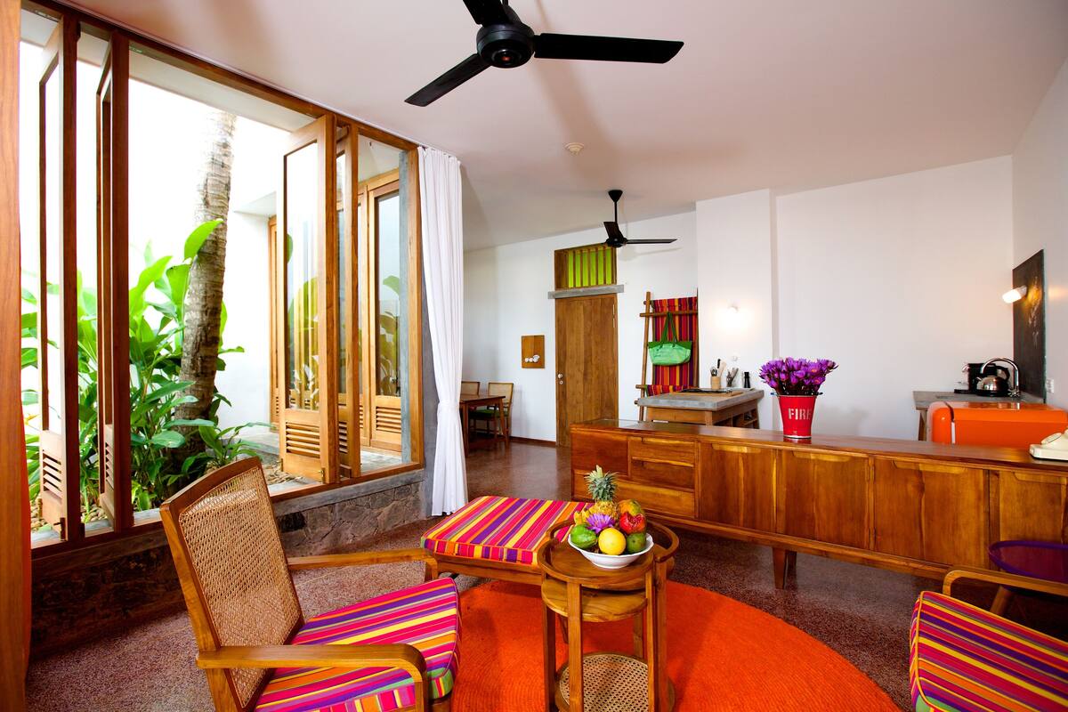 A spacious living area with a wooden bar, a dining table with colorful cushions, and a large window with curtains.