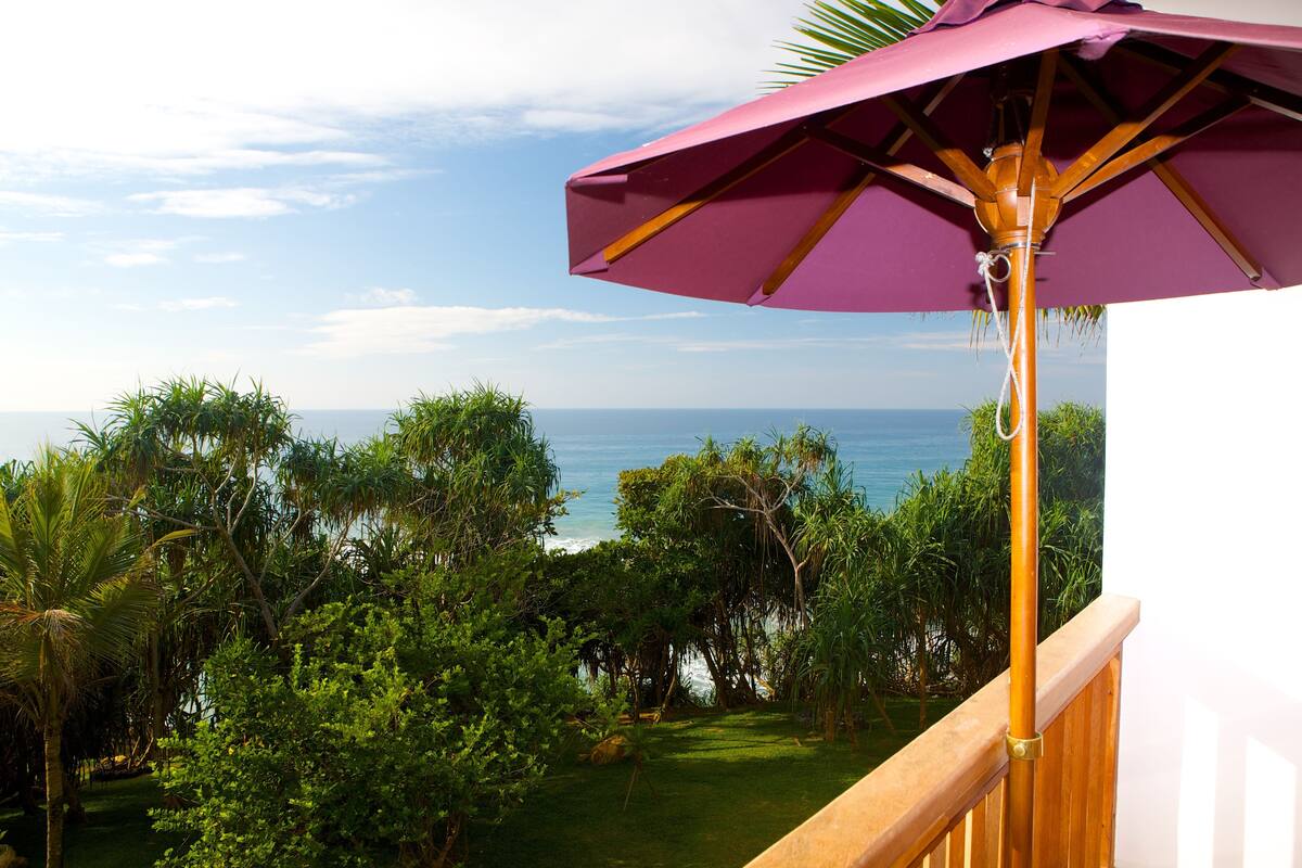 A balcony with a wooden railing overlooking a lush garden and the ocean.