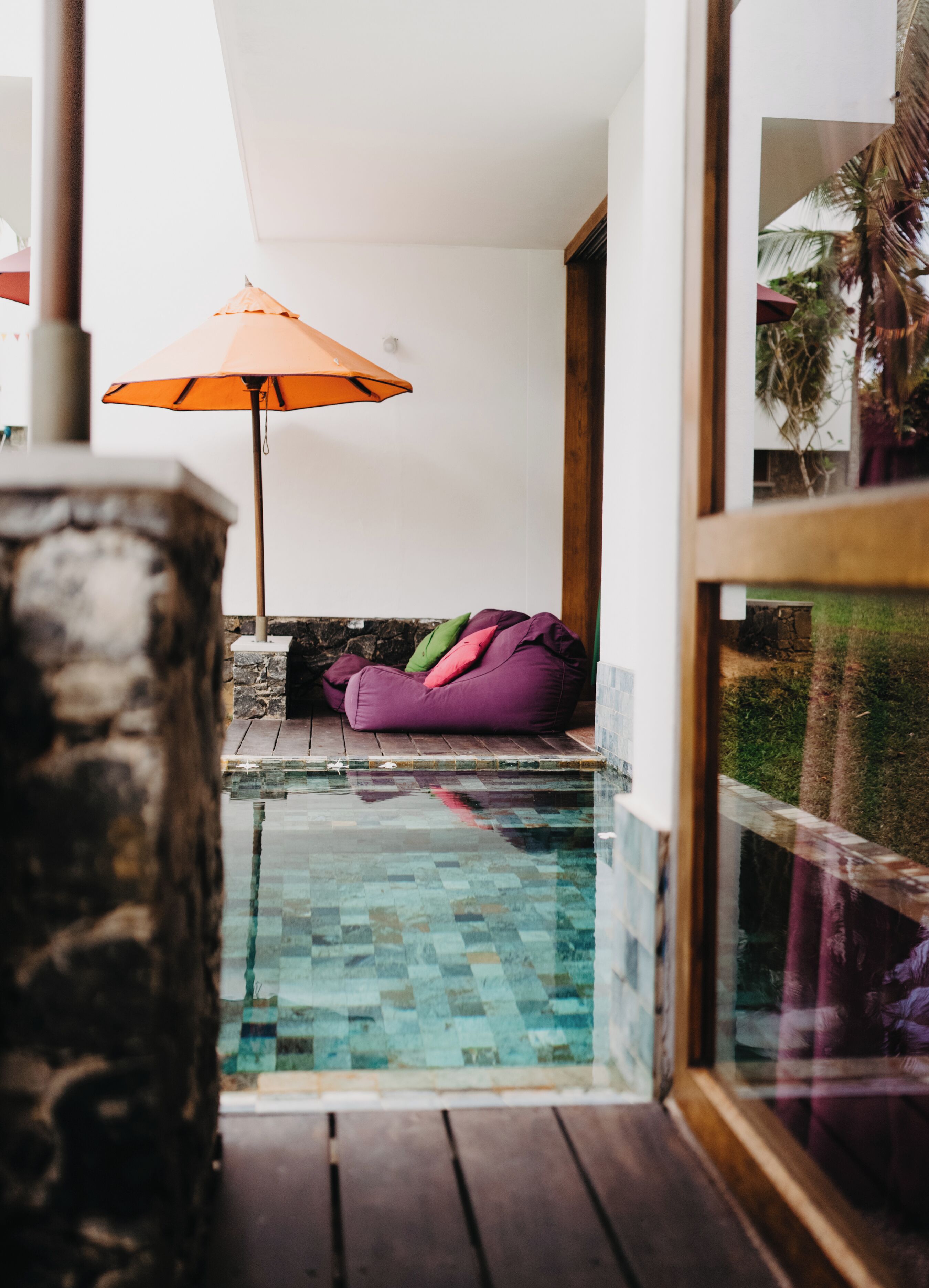 Un área de piscina con muro de piedra, terraza de madera, un sillón de saco morado y un paraguas naranja.