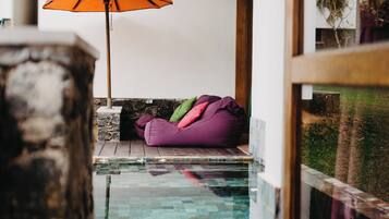 Un área de piscina con muro de piedra, terraza de madera, un sillón de saco morado y un paraguas naranja.
