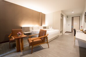 A modern hotel room with a bed, two wooden chairs, a small table with a vase of flowers, a desk with a lamp, and a bathroom visible through an open door.
