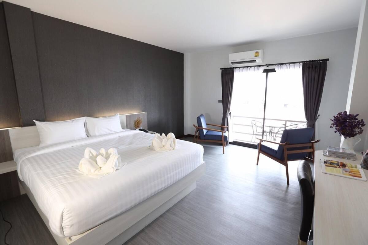 A hotel room with a large bed, a desk with a chair, a balcony with a chair, and an air conditioning unit.