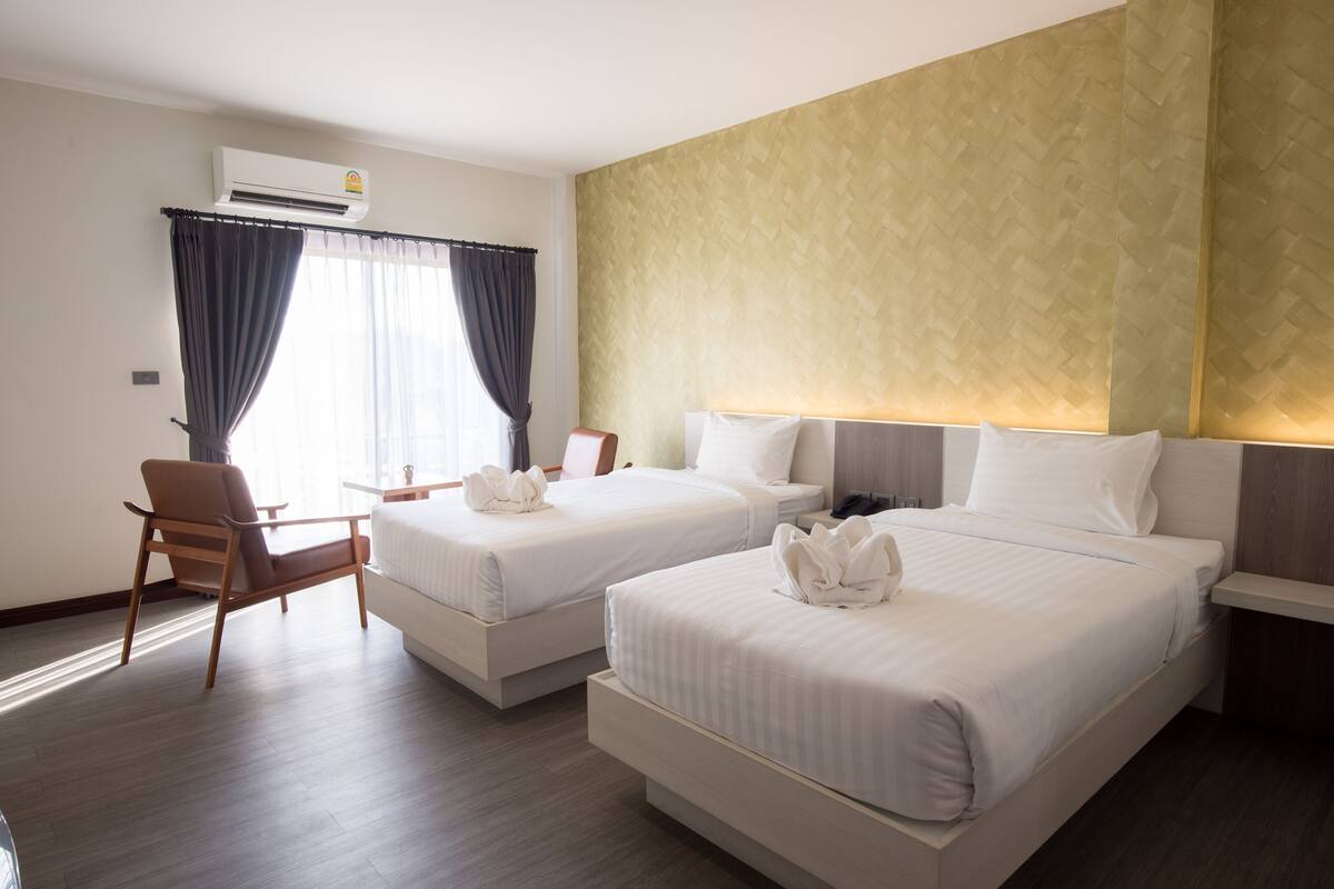 A hotel room with two beds, a chair, a small table, and an air conditioning unit.