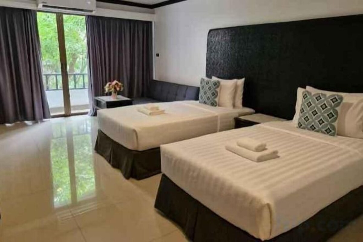 A hotel room with two beds, a nightstand, and a view of greenery outside.