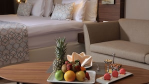 Una habitación de hotel con cama, sofá, mesa de comedor con fruta y postres, y mesita de noche con lámpara.