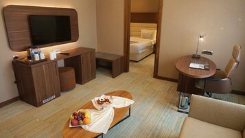 A hotel room with a TV, a desk with a computer, a bed, a small table with fruit, and a sofa.