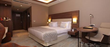 A hotel room with a large bed, a desk with a chair, a bedside table, and a lamp.