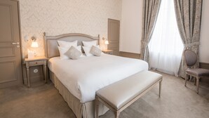 Un dormitorio con una cama grande, dos mesitas de noche con lámparas, un banco, una silla y una ventana con cortinas.