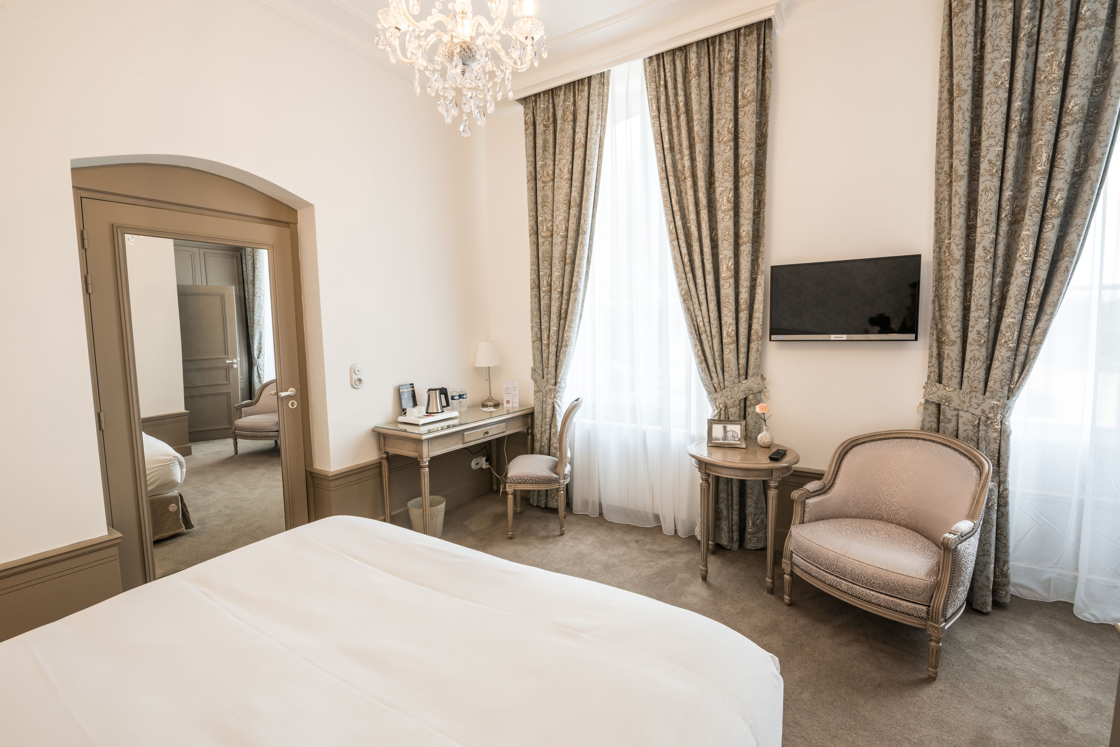 A hotel room with a large bed, a desk, a chair, a television, and a chandelier.