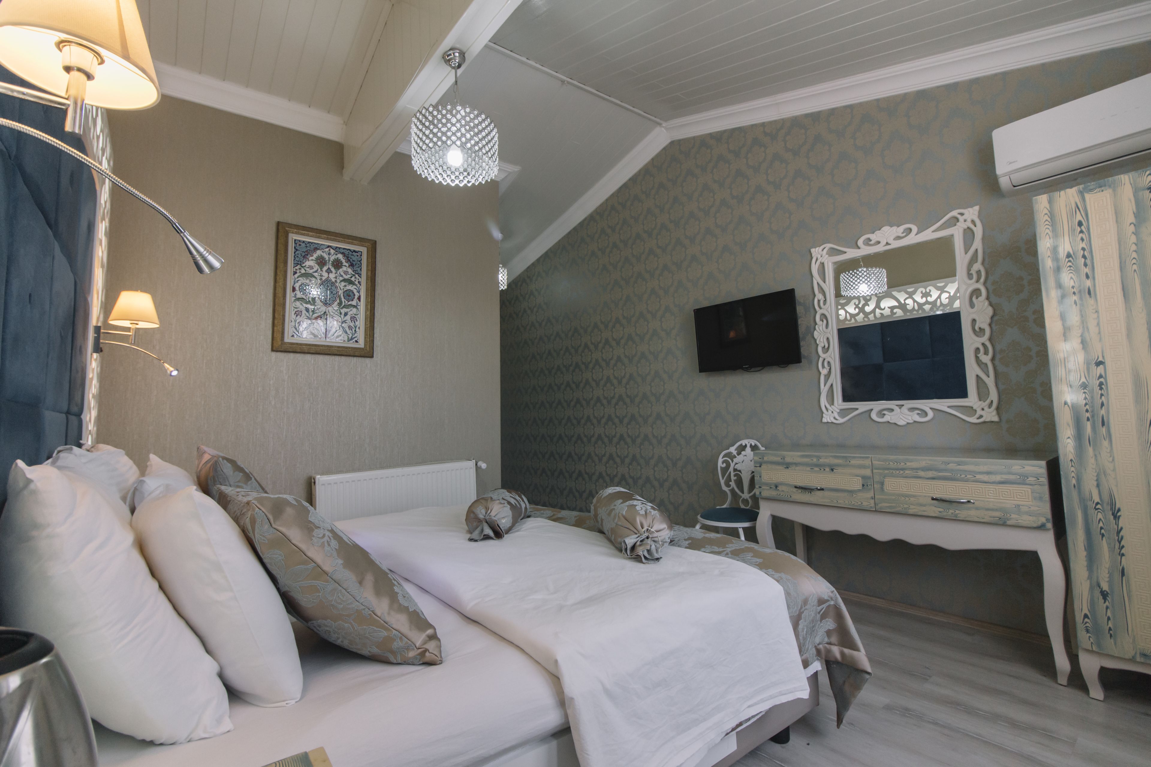 Una cama bien hecha con almohadas, una lámpara de noche, un espejo decorativo y un aparador con un televisor encima.