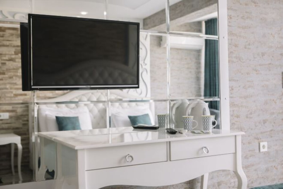 A modern bedroom with a bed, a TV mounted on a glass partition, a white dresser with drawers, and a bedside table with cups and a book.