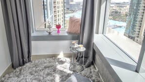 Une chambre d’hôtel moderne avec une grande fenêtre, un tapis moelleux, une petite table surmontée d’un vase rose, et une vue sur le paysage urbain et l’eau.