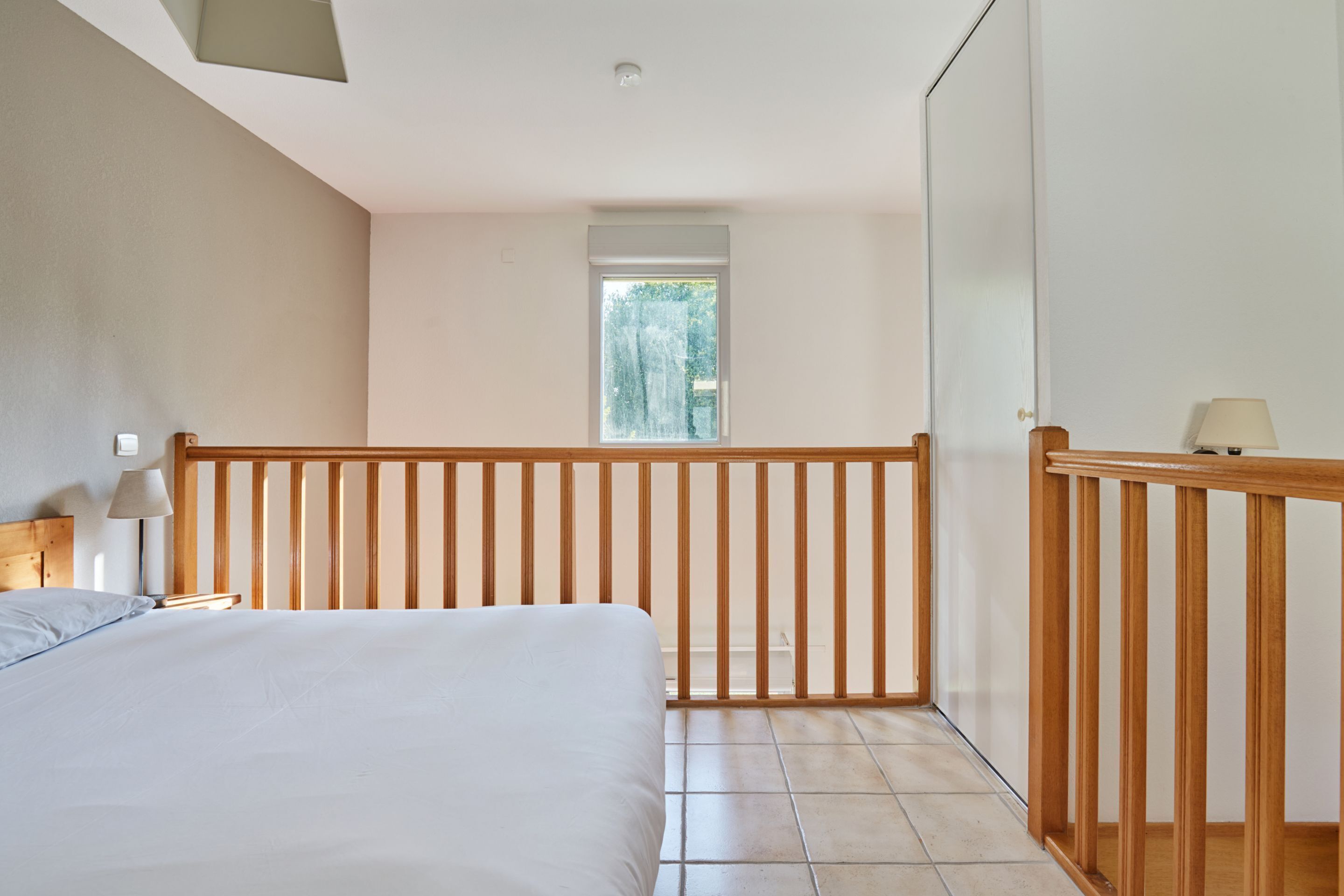 A bedroom with a large bed, a wooden railing, a window, and a nightstand with a lamp.