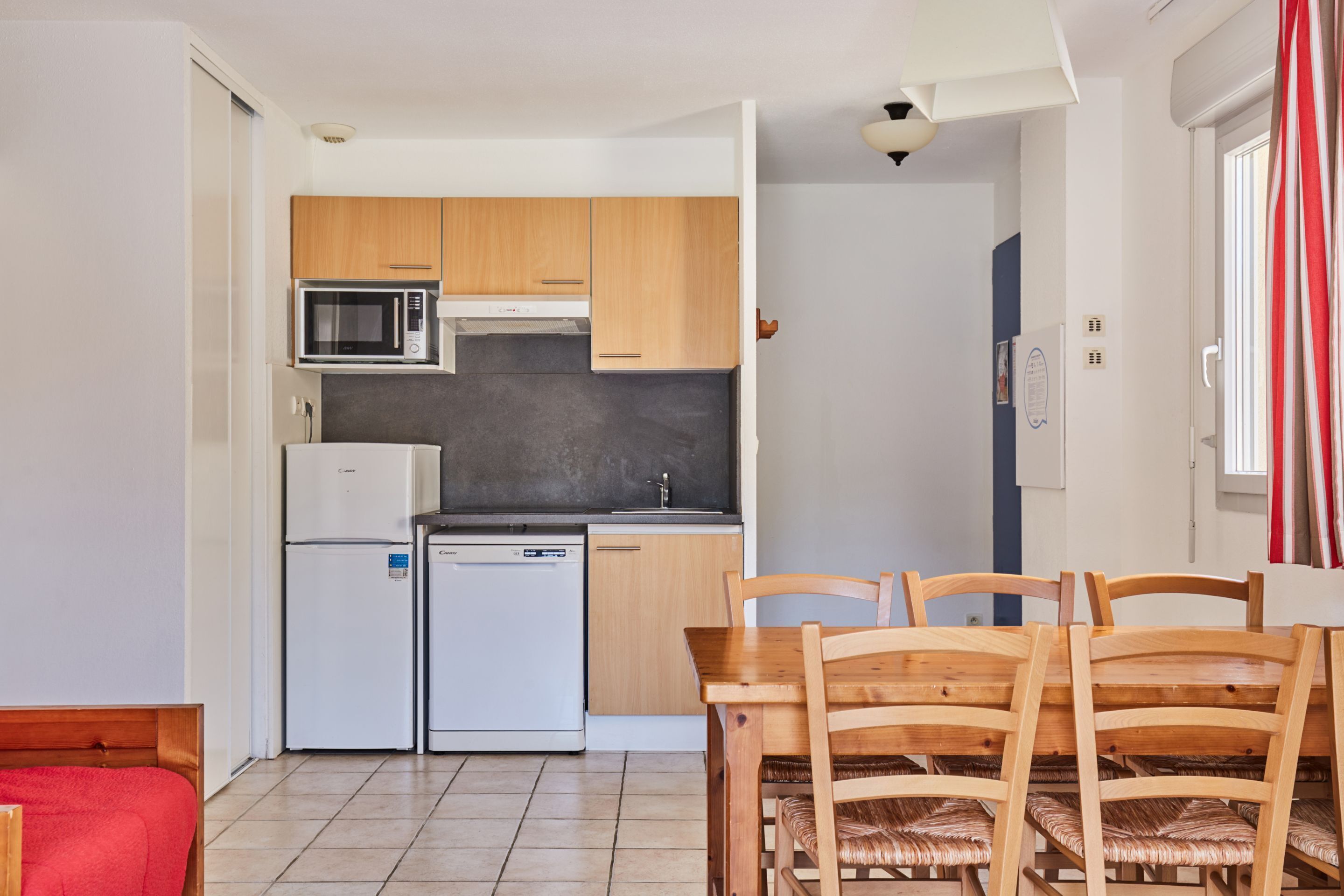 A compact kitchen with a microwave, refrigerator, and dishwasher, a wooden dining table with chairs, and a red sofa in the living area.
