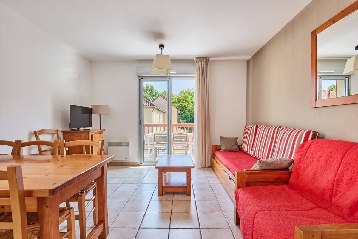 Ein Wohnzimmer mit einem Holzesstisch, Stühlen, einem roten Sofa, einem Fernseher und einer Glastür zum Außenbereich.