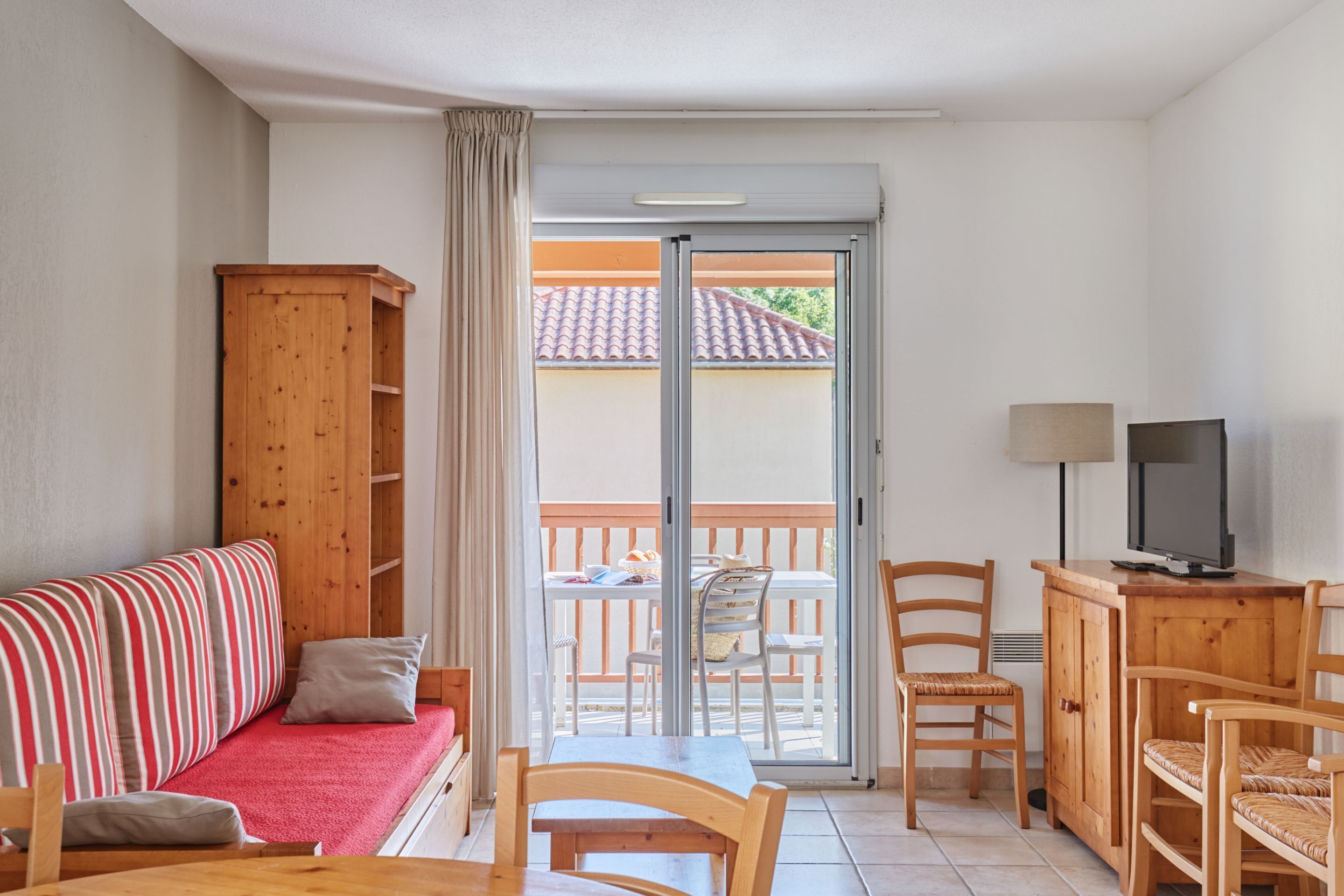 A room with a sofa, a dining table, chairs, a TV, and a sliding glass door leading to an outdoor area with a table and chairs.