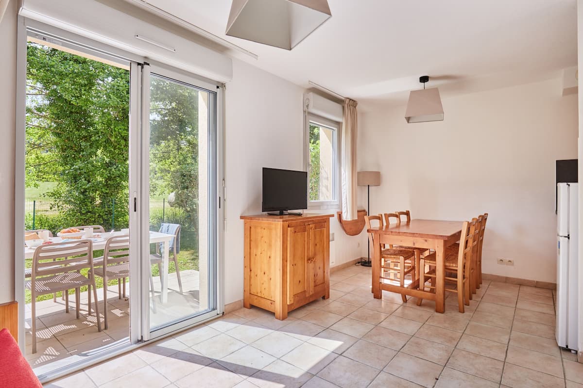 Ein geräumiges Zimmer mit einer Schiebeglastür, die zu einem Außenbereich führt, einem Holzessttisch mit Stühlen, einem Fernseher auf einer Holzkommode und einem weißen Kühlschrank.