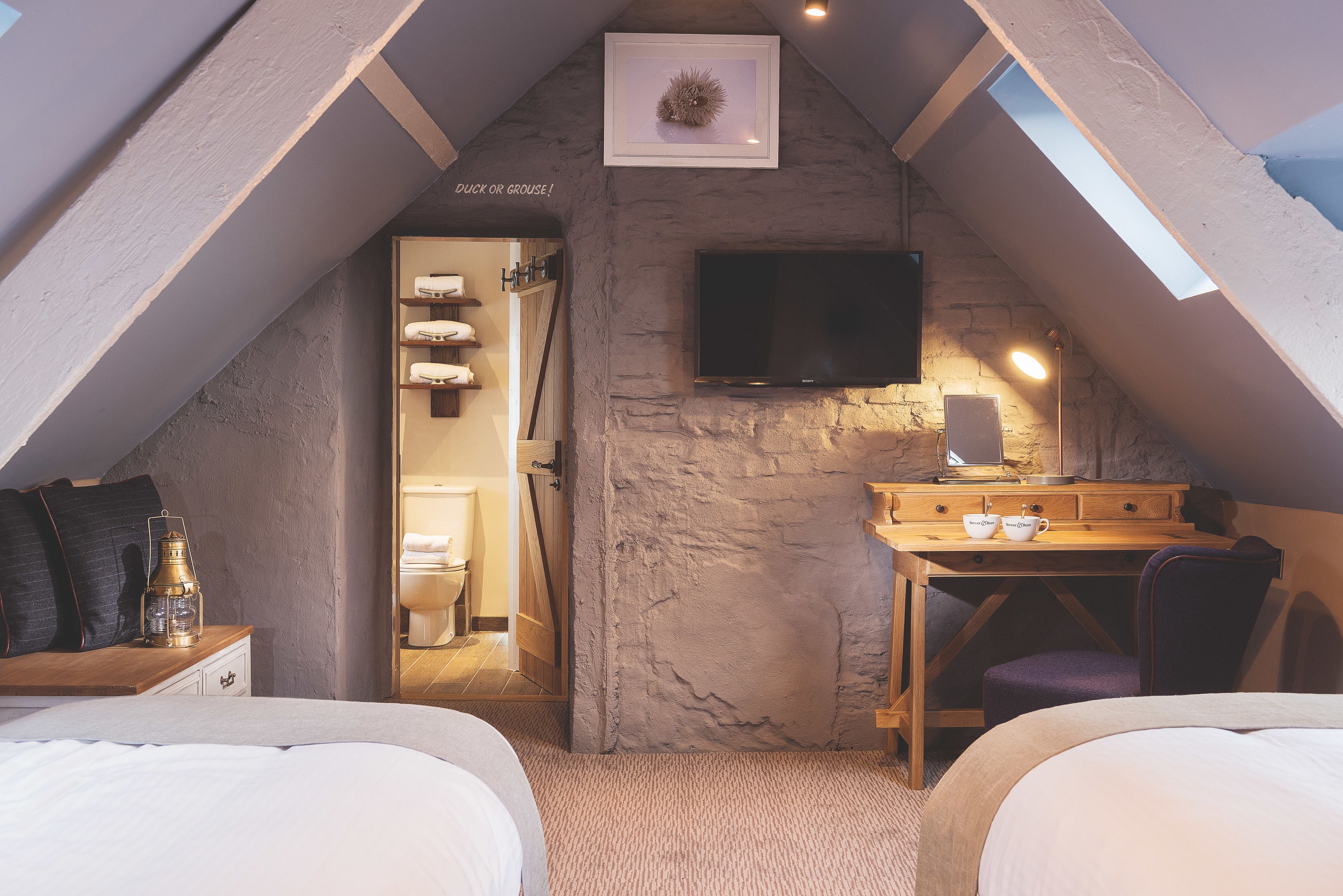 Une chambre confortable située sous les toits, avec un plafond en pente, un téléviseur fixé au mur, un bureau avec une chaise et un lit avec une table de chevet.