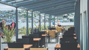 An outdoor café with wooden tables and wicker chairs, overlooking a harbor with boats and buildings in the background. - Haveners (Fowey)