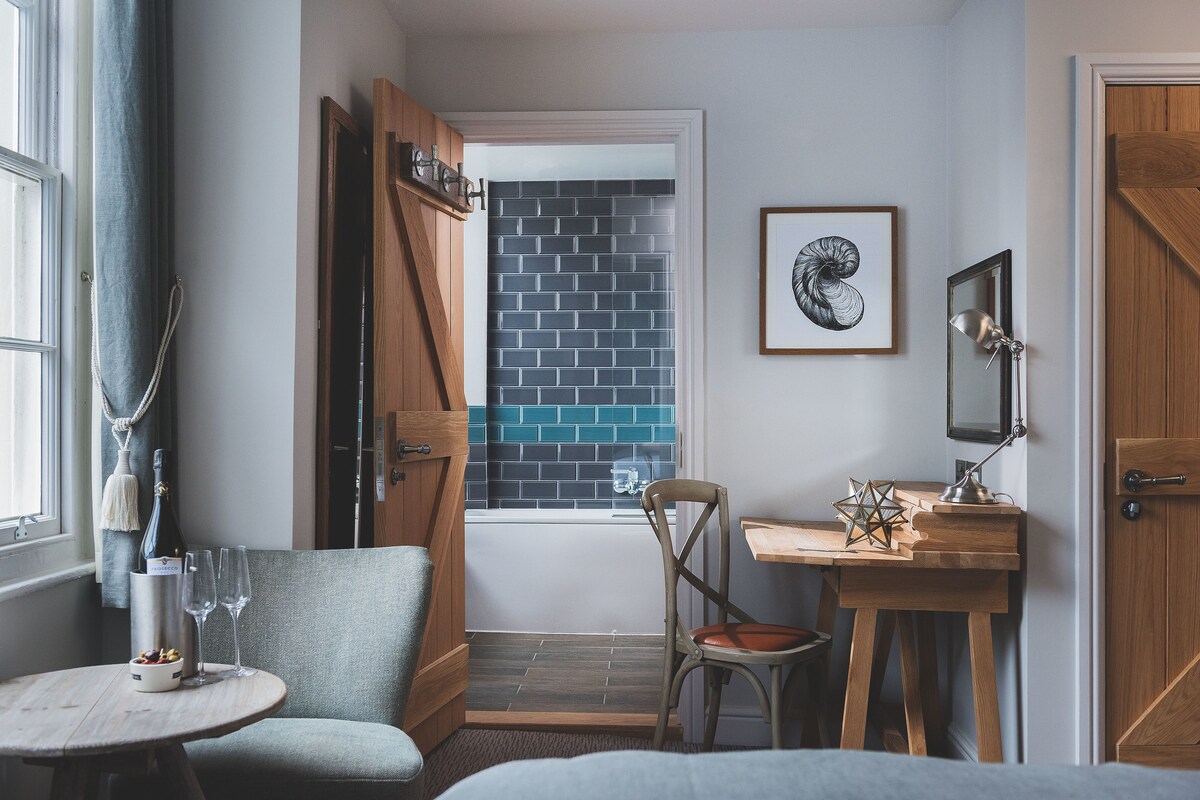 Een kamer met een houten bureau, een stoel, een klein tafeltje met een fles en glazen, een lijst met een foto, en zicht op een blauw betegelde muur door een geopende deur.