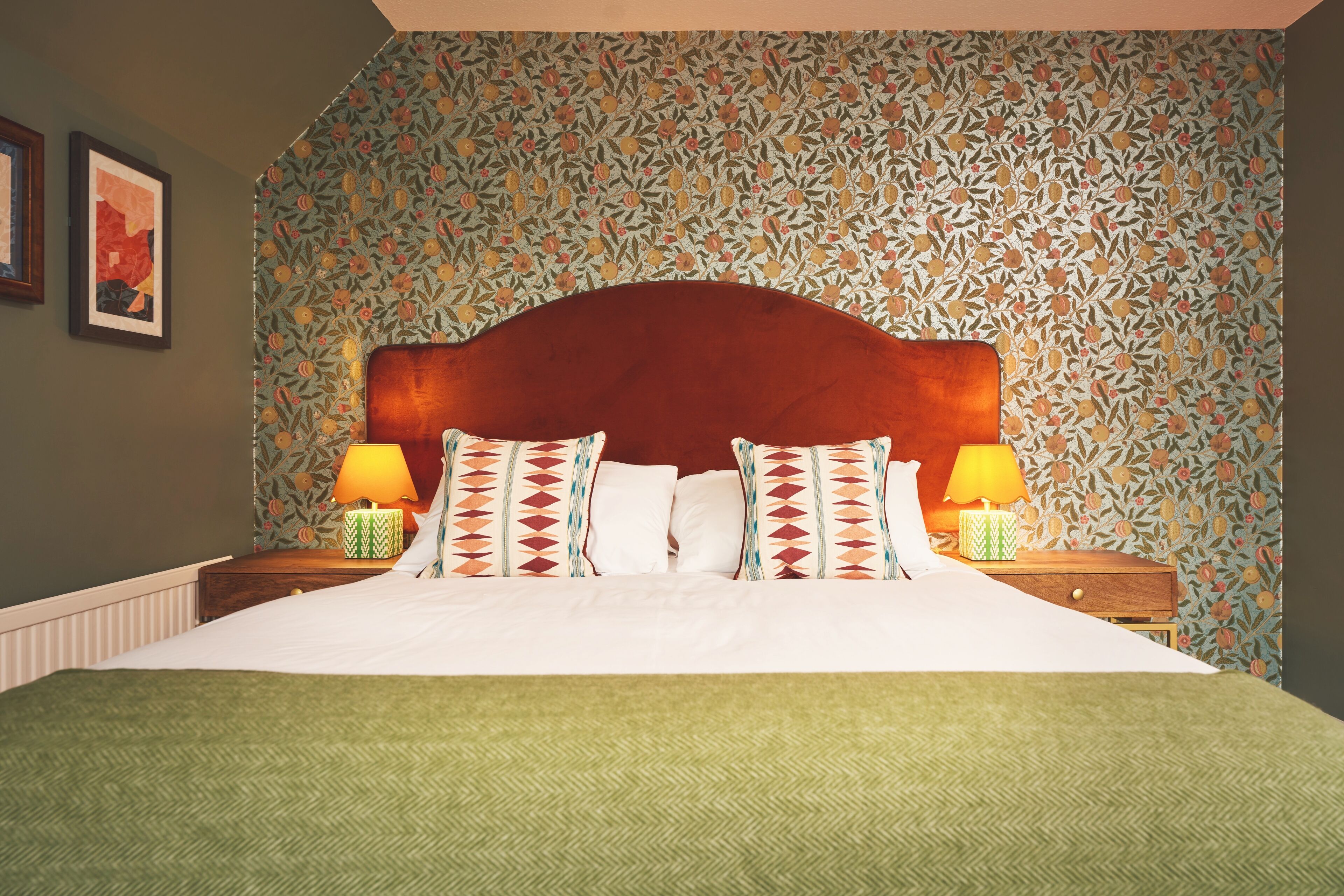 Cama bem arrumada com colcha estampada em padrão herringbone, duas luminárias de cabeceira e papel de parede floral.
