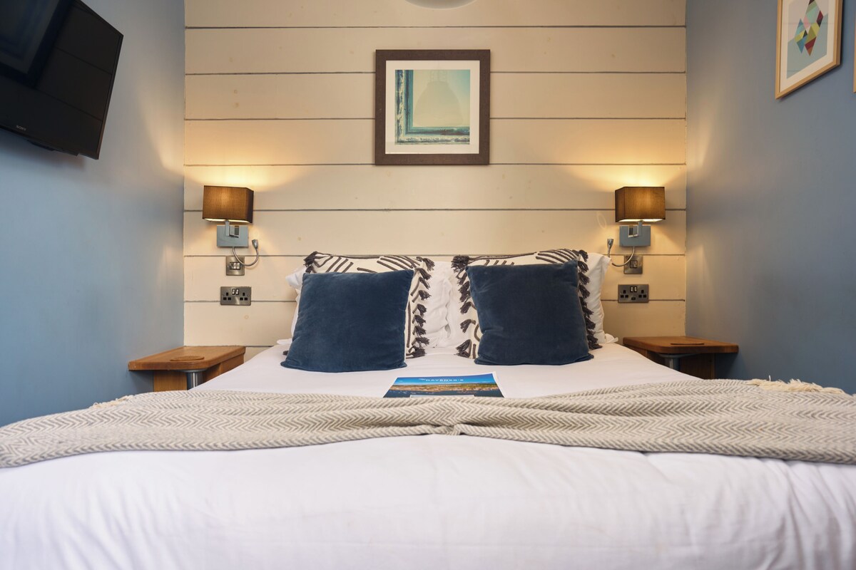Een net opgemaakt bed met een bedsprei met patroon, twee kussens en een dekbed. Twee nachtkastjes met lampen, een aan de muur bevestigde televisie en een lijst met een afbeelding boven het bed.