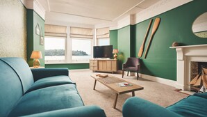 A living room with a green wall, a wooden coffee table, a blue sofa, a television, and a fireplace. - Haveners (Fowey)