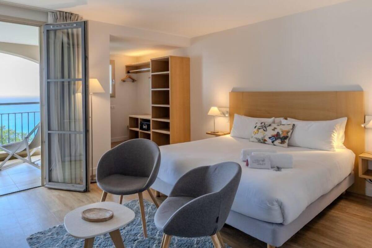 A hotel room with a large bed, two armchairs, a small table, a wardrobe, and a balcony with a view.