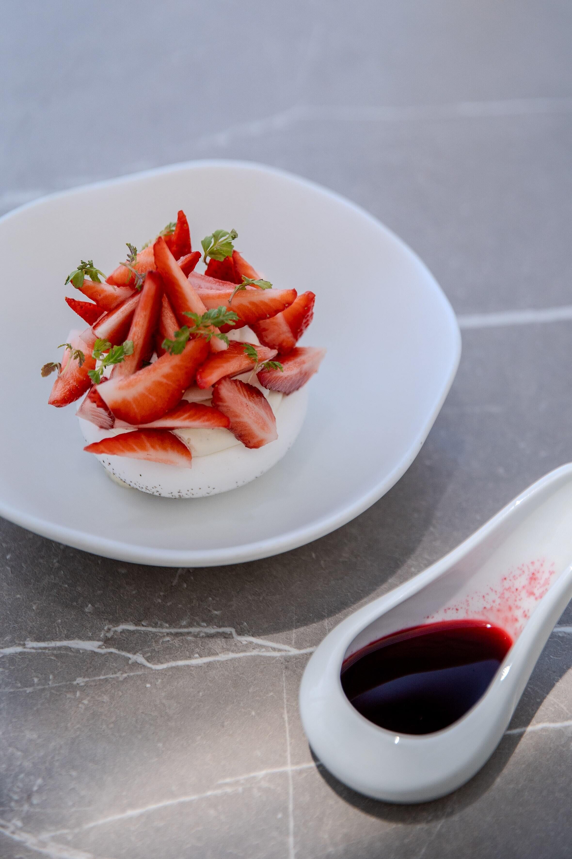 A dessert on a white plate with a white spoon containing red sauce beside it.