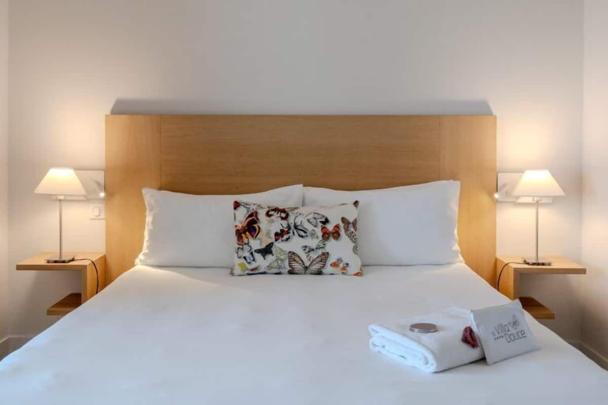 A hotel room with a wooden headboard, white bedding, a floral pillow, two bedside lamps, and a neatly folded towel on the bed.