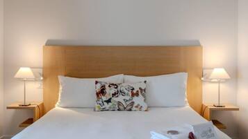 Habitación de hotel con cabecera de madera, ropa de cama blanca, un cojín floral, dos lámparas de noche y una toalla doblada con cuidado sobre la cama.