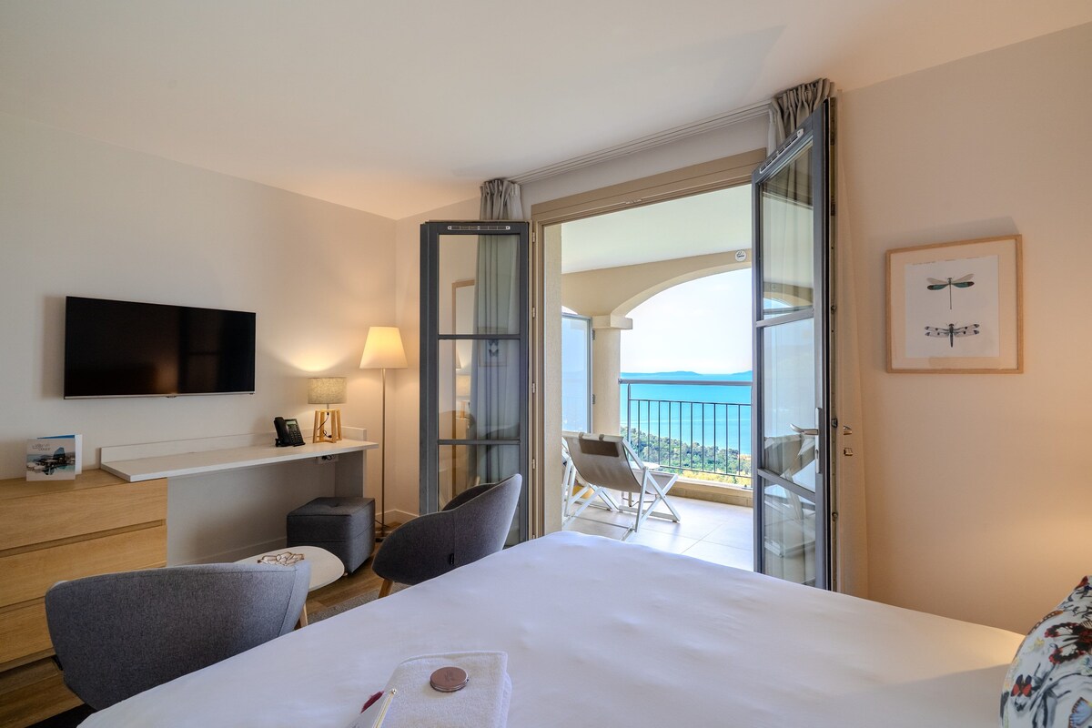 A hotel room with a bed, a TV, a desk, a chair, a balcony with a view, and a framed picture on the wall.