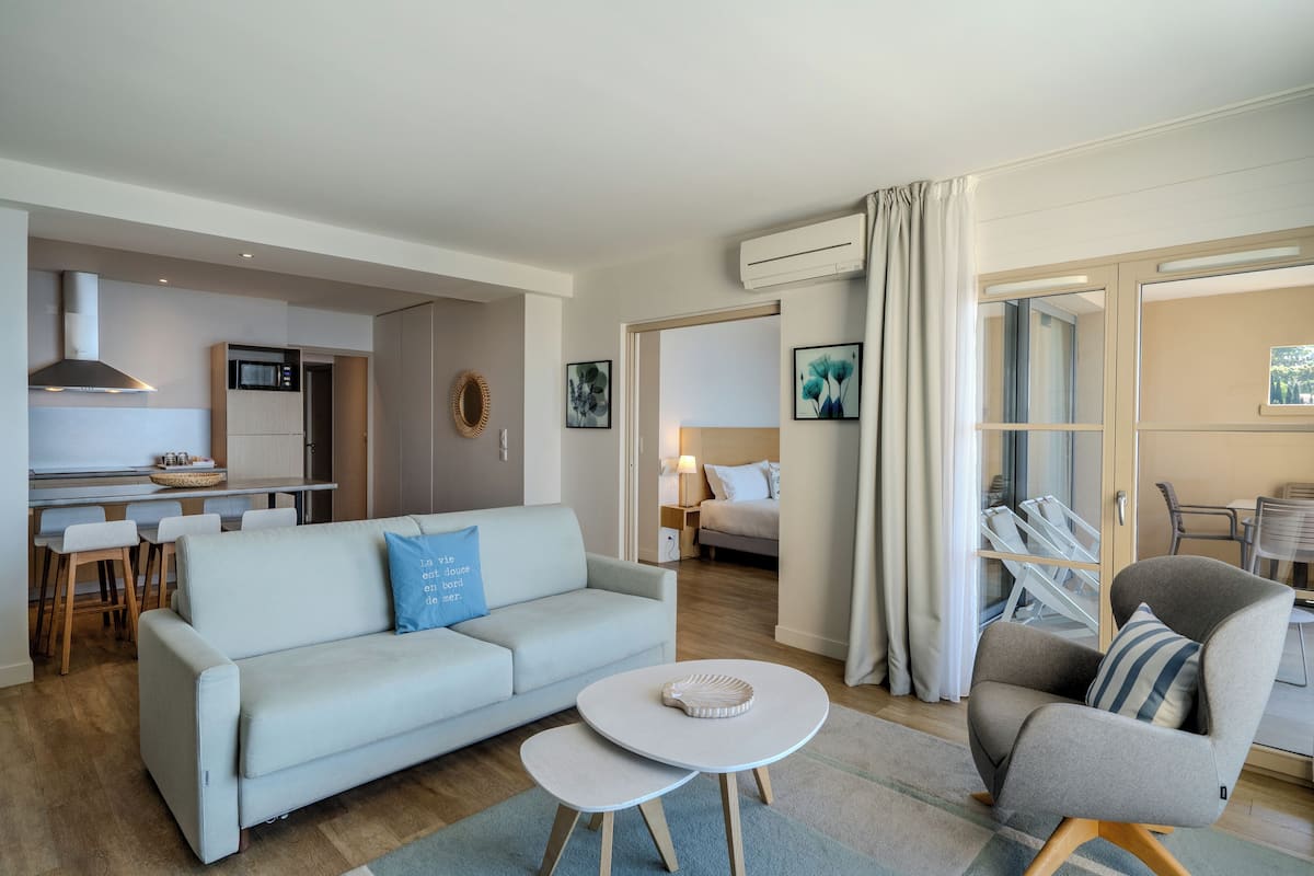 A modern living room with a sofa, armchair, and coffee table, a kitchen area with a dining set, and a bedroom with a bed and nightstand.