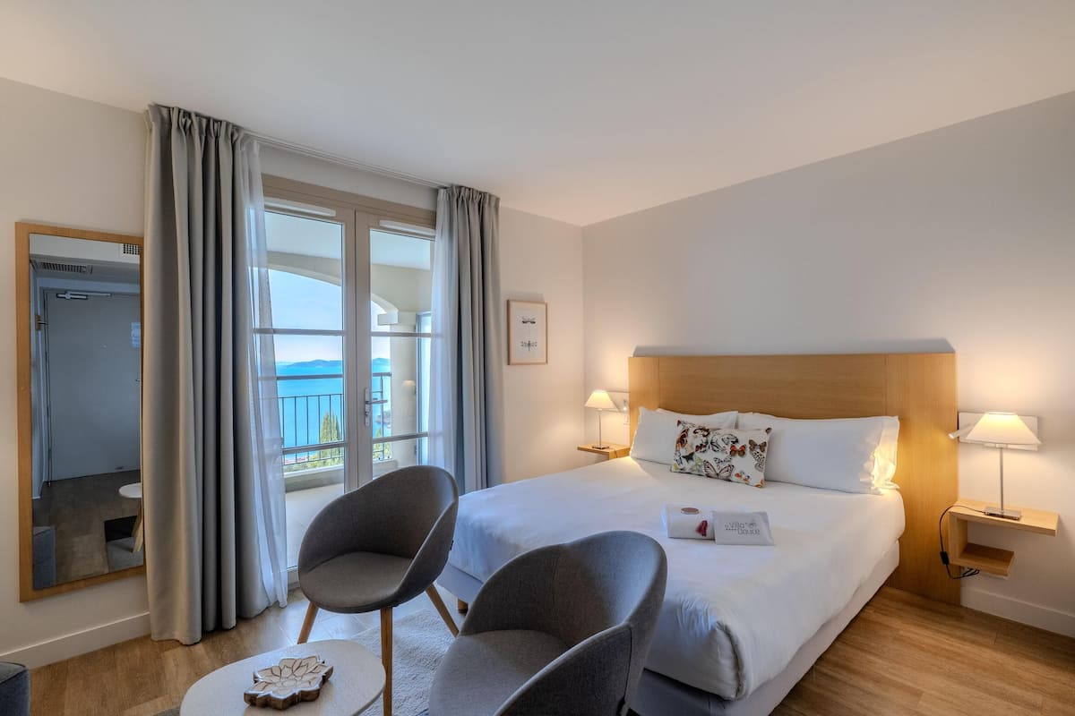 A hotel room with a bed, two armchairs, a bedside table, a lamp, a mirror, and a view of the outdoors.