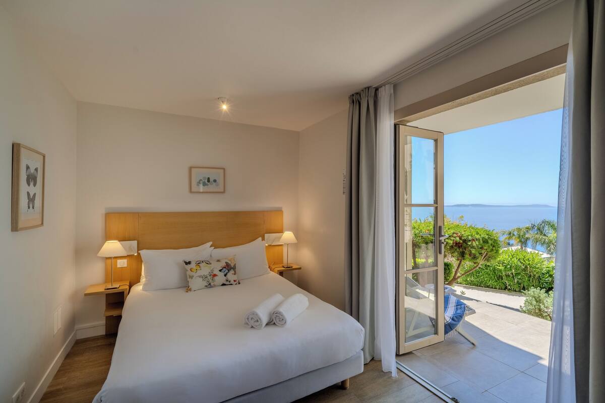A hotel room with a large bed, two bedside tables with lamps, a view of the outdoors, and a balcony.