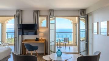 Een moderne kamer met een flatscreen-tv, een bureau, twee stoelen, een klein tafeltje en uitzicht op de zee door grote glazen deuren.