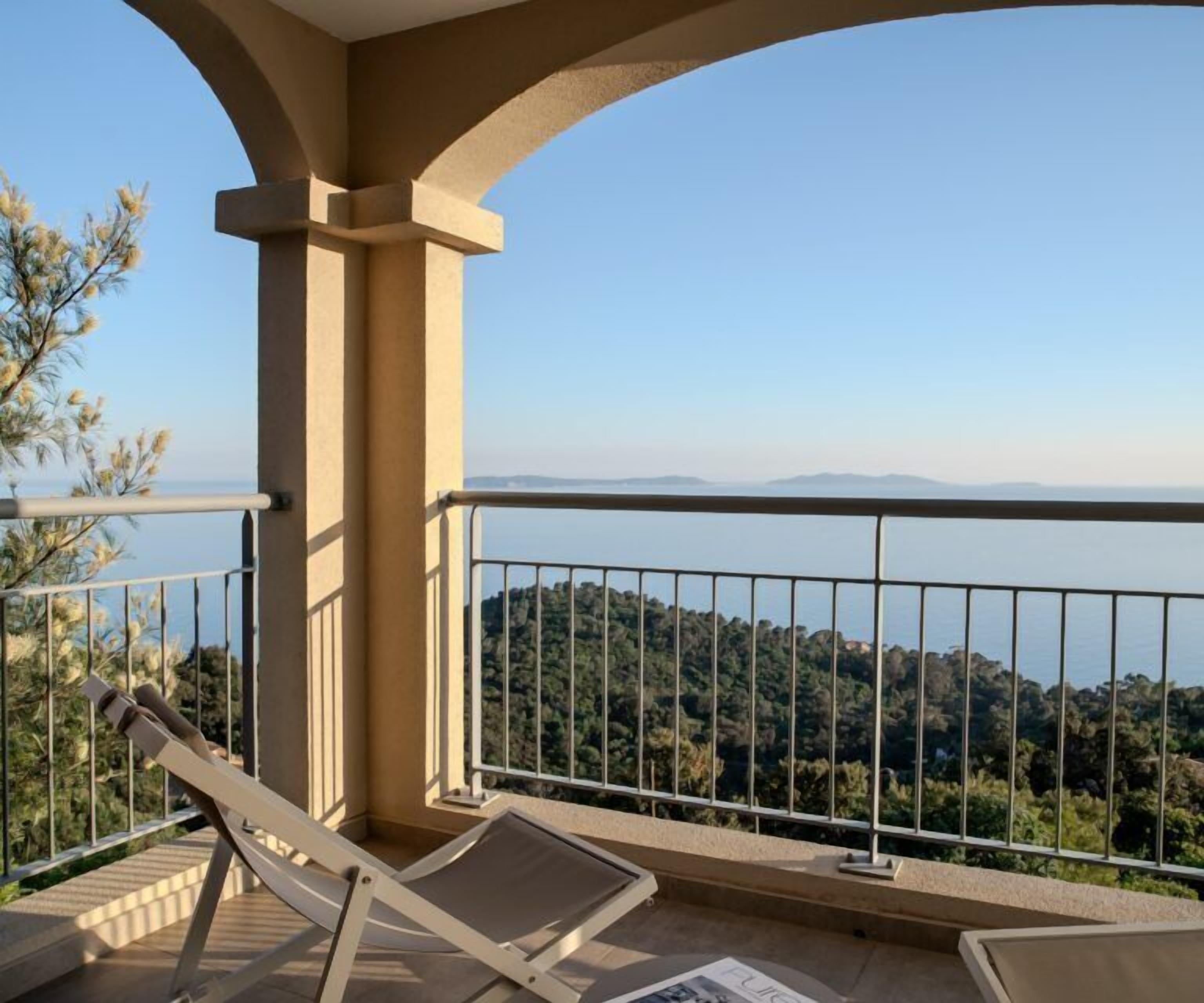 A balcony with a view of the sea and hills, a lounge chair, and a railing.