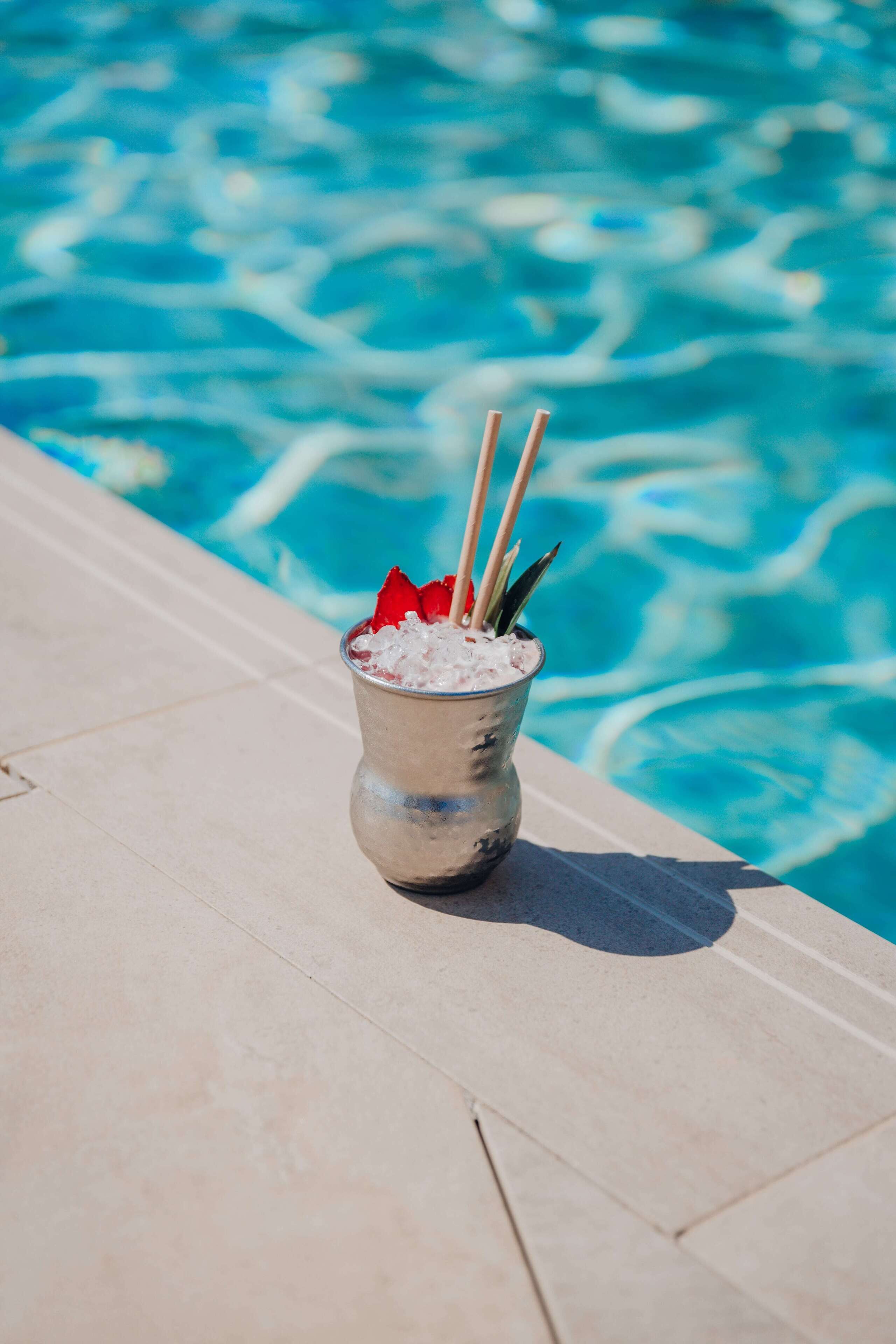 A drink with a straw and a red garnish in a metal cup on a poolside.