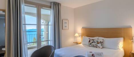 A hotel room with a bed, two armchairs, a bedside table, a lamp, and a view of the sea through large windows.